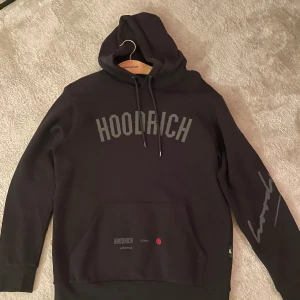 Svart hoodie från Hoodrich - Säljer en svart hoodie från Hoodrich med tryck på bröstet och ärmen. Den har en stor ficka framtill och justerbar huva med snören. Perfekt för en avslappnad stil.