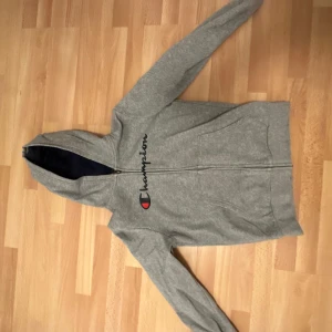 Grå hoodie från Champion - Säljer en grå hoodie från Champion med dragkedja och deras ikoniska logga på bröstet. Tröjan har långa ärmar och en praktisk huva. Perfekt för en avslappnad stil.