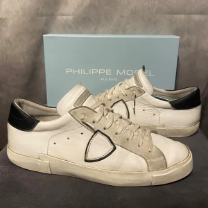 Philippe Model  - Snygga vita sneakers från Philippe Model med svarta detaljer och snörning. Skorna har en stilren design med en diskret logga på sidan och en bekväm passform. Perfekta för en avslappnad stil. 