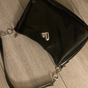 Svart axelväska med hjärtdetalj - Snygg svart axelväska i glansigt material med en stilren hjärtdetalj i silver på framsidan. Väskan har en praktisk dragkedja och en avtagbar axelrem. Perfekt för att ge en elegant touch till din outfit.