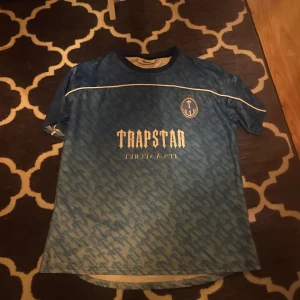 Blå t-shirt från Trapstar - Säljer en blå t-shirt från Trapstar med ett unikt mönster och tryck på både fram- och baksidan. T-shirten har korta ärmar och en rund halsringning. Perfekt för en avslappnad stil. Storlek S men passar även M minst lika bra;)