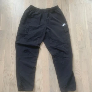 Svarta cargobyxor från Nike - Säljer ett par svarta cargobyxor från Nike med elastisk midja och praktiska sidofickor. Byxorna har en avslappnad passform och är perfekta för en sportig look. Nike-loggan syns tydligt på ena benet.