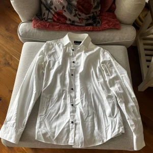 Vit skjorta från Riley - Säljer en stilren vit skjorta från Riley i slim fit. Skjortan har långa ärmar och svarta knappar som ger en snygg kontrast. Perfekt för en elegant look.