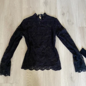 Svart spetsblus från Vintage Stories - Elegant svart spetsblus från Vintage Stories med långa ärmar och vacker spetsdetaljering. Perfekt för en stilfull look med en feminin touch. Blusen har en hög hals och fina kanter.