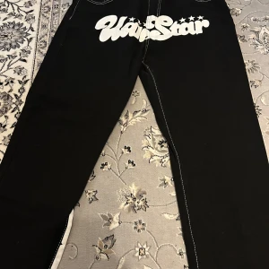 Svarta jeans med vit text - Coola svarta jeans med vit text 'Hardstar' tryckt över baksidan. De har kontrasterande vita sömmar och en läderlapp med logotypen på baksidan. Perfekta för en avslappnad stil. Säljs pågrund av dem blivit för stora på mig. Pris kan diskuteras!