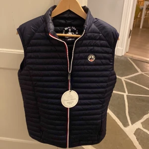 Svart dunväst från Jott - Snygg svart dunväst från Jott med dragkedja och ett stilrent märke på bröstet. Perfekt för lager-på-lager under kyliga dagar. Västen har en quiltad design och en smidig passform.