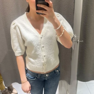 Vit kofta med knappar - Säljer en söt vit kofta med v-ringning och knappar framtill. Koftan har korta ärmar och en mjuk, bekväm passform. Perfekt för att lägga till en touch av elegans till din outfit. Passar bra till både jeans och kjol.