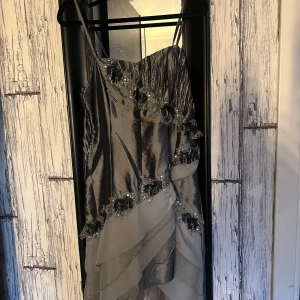 Silverfärgad klänning med blommiga detaljer - Elegant silverfärgad klänning med smala axelband och blommiga detaljer. Klänningen har en omlott-design med volanger och en smockad rygg för en bekväm passform. Perfekt för speciella tillfällen.