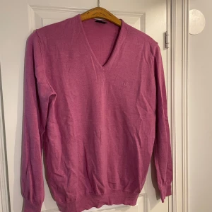 Rosa tröja från Oscar Jacobson - Säljer en stilren rosa tröja från Oscar Jacobson med v-ringning. Tröjan har långa ärmar och ett diskret broderat märke på bröstet. Perfekt för en avslappnad men ändå elegant look.