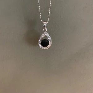 Silverhalsband med svart droppformad hänge - Elegant silverhalsband med ett droppformat hänge. Hänget har en svart sten i mitten omgiven av glittrande stenar. Kedjan är fin och kulformad, vilket ger en stilren look. Perfekt för att ge en touch av elegans till din outfit. Kedja: ca 40 cm. Berlock: ca 1,5 cm x 2 cm. Säljer då det inte kommer till användning. 