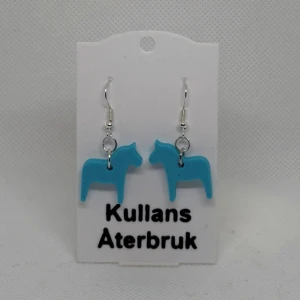 Blå dalahäst örhängen från Kullans Återbruk - Charmiga örhängen i form av blå dalahästar från Kullans Återbruk. Perfekta för att lägga till en unik touch till din stil. Tillverkade med omsorg och kreativitet.