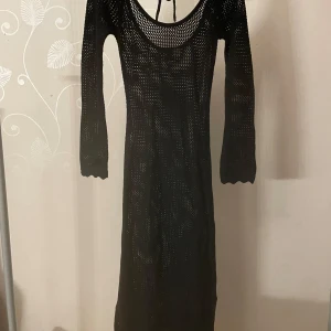 Svart stickad långklänning - Snygg svart stickad långklänning med långärmad design och djup urringning. Perfekt för en stilren look med en touch av elegans. Klänningen har en öppen rygg med knytband för extra detalj.