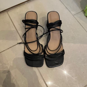 Svarta klackade sandaler med snörning - Snygga svarta sandaler med hög klack och elegant snörning runt ankeln. De har en fyrkantig tå och är perfekta för att ge en stilren look. Tillverkade i syntetmaterial.