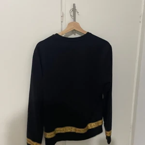 Svart tröja från EA7 Emporio Armani - Snygg svart tröja från EA7 Emporio Armani med gulddetaljer. Tröjan har en liten logga på bröstet och guldfärgad text vid ärmslut och nederkant. Perfekt för en stilren look.