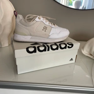 Tommy Hilfiger sneackers - Snygga beige sneakers med vit sula och snörning. Skorna har en diskret logga på sidan och en dragflik baktill för enkel påtagning. Perfekta för en stilren och avslappnad look.