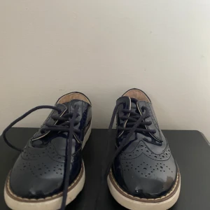 Svarta lackskor med snörning - Snygga svarta lackskor med snörning och dekorativa perforeringar. Skorna har en vit sula som ger en modern touch. Perfekta för en stilren look.