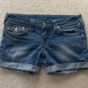 Jeansshorts från True Religion - Snygga jeansshorts från True Religion i en klassisk blå färg. De har en lågmidjad design med uppvikta ben och dekorativa knappar på bakfickorna. Perfekta för en avslappnad stil.