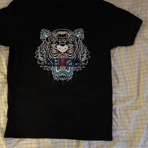 Svart t-shirt med tigertryck från Kenzo - Säljer en svart t-shirt från Kenzo med ett stort tigertryck i flera färger på framsidan. T-shirten har en klassisk rund hals och korta ärmar. Perfekt för dig som vill ha en stilren men ändå iögonfallande look. Pris kan diskuteras 