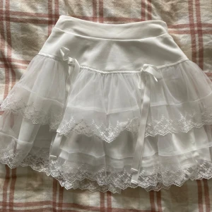 Vit tyllkjol med spetsdetaljer - Charmig vit tyllkjol med flera lager och spetsdetaljer. Kjolen har en elastisk midja och dekorativa rosetter på sidorna. Aldrig använd, Priset går att diskuteras! 💗