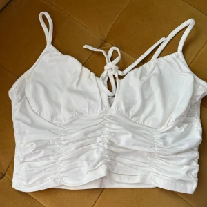 Vit croppad linnetopp från NLY Trend - Supersöt vit croppad linnetopp från NLY Trend med smala axelband, knytning framtill och rynkade detaljer över magen. Toppen har en lätt v-ringning och är tillverkad i mjuk bomull, perfekt för varma dagar och snygga outfits.