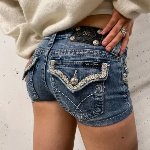 Miss Me jeansshorts med paljettdetaljer - Snygga blå jeansshorts från Miss Me med lågt skuren midja. Shortsen har blingade paljettdetaljer och silvriga nitar på fickorna, samt dekorativa sömmar och broderier. Perfekta för dig som vill sticka ut med en edgy och glam look i sommar.
