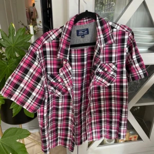 Rutig rosa skjorta från Skill, M - Rutig skjorta i rosa, svart och vitt från Skill. Skjortan har korta ärmar, klassisk krage, två bröstfickor med knappar och en avslappnad passform. Perfekt att bära öppen över ett linne eller knäppt för en cool look.