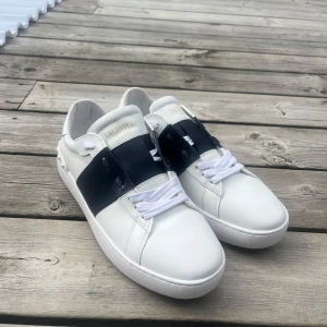Valentino Garavani vita sneakers - Snygga vita Valentino Garavani sneakers i skinn med bred svart rem över vristen och vita snören. Klassisk rund tå och platt sula med coola nitar baktill. Perfekta för dig som vill ha en stilren men ändå unik look.