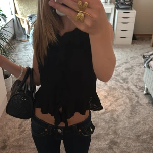 Svart volangblus från Gina Tricot - Supersöt svart blus från Gina Tricot med volanger längs hela framsidan och nertill. Ärmlös modell med lätt skrynklig struktur. Perfekt för sommaren och är använd ett fåtal gånger. 