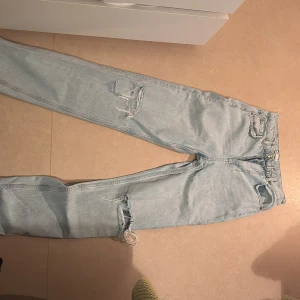 Ljusblå ripped jeans från Perfect Jeans - Säljer ett par ljusblå jeans från Perfect Jeans med snygga slitningar på benen. Klassisk femficksmodell, hög midja och raka ben. Jeansen har dragkedja och knapp framtill. Perfekta för en avslappnad och trendig look.
