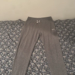 Grå mjukisbyxor från Hugo Boss - Grå mjukisbyxor från Hugo Boss med vit snörning i midjan och diskret logga på vänster lår. Sköna och avslappnade, perfekta för chill eller träning. Tillverkade i mjukt bomullsmaterial för extra komfort.