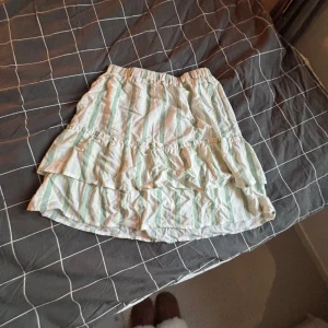 Randig volangkjol i pastell - Söt kortkjol med volanger och pastellfärgade ränder i vitt, ljusgrönt och ljusgult. Kjolen har resår i midjan och är perfekt för en somrig look. Lätt och luftig, passar dig som gillar en lekfull stil. Stor i storleken, passar upp till xs/s.