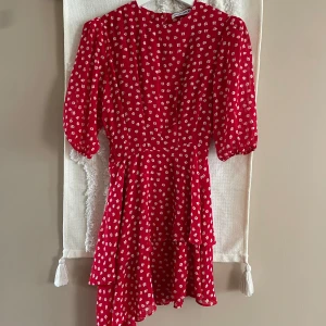 Röd blommig kortklänning från Adoore - Supersöt röd kortklänning från Adoore med små vita blommor, volangdetaljer och puffiga korta ärmar. Klänningen har en figurnära passform i midjan och är tillverkad i 100% polyester. Perfekt för sommarens alla tillfällen!