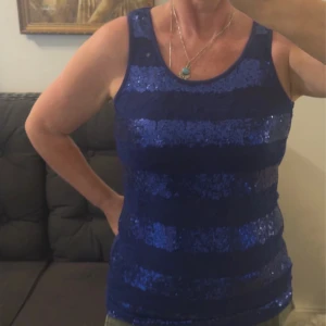 Blå paljettlinne från MICHA - Snyggt blått linne från MICHA med breda paljettband i horisontella ränder och spetsdetaljer på framsidan. Ärmlös modell med rund halsringning och mjukt innerfoder. Perfekt för dig som vill sticka ut med lite extra glitter.