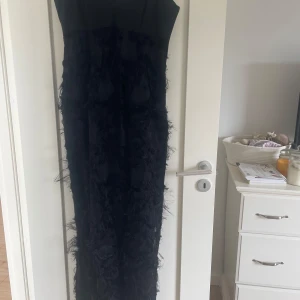 Svart långklänning Tiger of Sweden - Supersnygg svart långklänning från Tiger of Sweden med v-ringad överdel och coola fjäderliknande detaljer över hela kjolen. Klänningen har en elegant och smal siluett, perfekt för dig som vill sticka ut med en unik look.