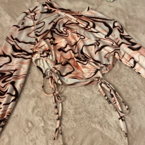 Swirlmönstrad croppad topp med snörning - Trendig långärmad croppad topp med swirlmönster i rosa, svart och beige. Toppen har snygga snörningsdetaljer på sidorna som gör att du kan justera passformen. Tillverkad i ett stretchigt syntetmaterial som sitter skönt på kroppen.