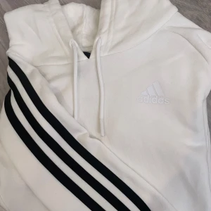 Vit hoodie från Adidas med svarta ränder - Snygg vit hoodie från Adidas med klassiska svarta ränder längs ena sidan och broderad logga på bröstet. Tröjan har huva med snörning och är perfekt för en sportig och avslappnad stil. Pris kan diskuteras. 
