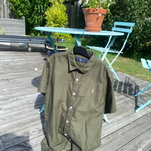 Olivgrön kortärmad skjorta Ralph Lauren - Snygg olivgrön skjorta från Ralph Lauren med klassisk krage och korta ärmar. Skjortan har vita knappar framtill och den ikoniska broderade Polo-loggan på bröstet. Tillverkad i bomull, perfekt för varma dagar och en avslappnad stil.