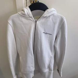 Vit hoodie från Sail Racing med dragkedja - Vit hoodie från sail racing med broderad logga på bröstet och en cool detalj på huvan. Tröjan har hel dragkedja, fickor på sidorna och är tillverkad i mjuk bomull. Perfekt för dig som gillar streetwear och enkel stil. Pris kan diskuteras. 