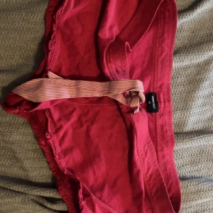 Rosa shorts med randigt knytband - Snygga rosa shorts med ett randigt knytband i midjan. Modellen är loose fit och har en bekväm passform. Perfekta för varma sommardagar och chill häng. Shortsen har en enkel design med en liten volangdetalj längs sidorna.