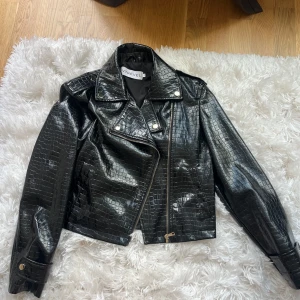 Svart croppad skinnjacka Ivyrevel - Säljer en svart croppad skinnjacka från Ivyrevel med snyggt krokopräglat mönster. Jackan har guldiga knappar, dragkedja och klassisk bikerkrage. 