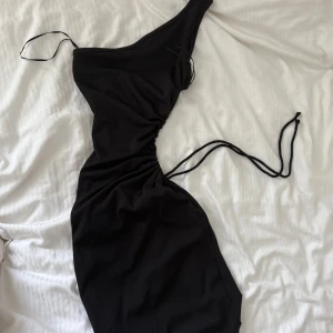 Svart one shoulder långklänning - Svart kortklänning one shoulder-design och smala axelband. Klänningenär lite öppen vid magen och har snygg snörning i sidan och rynkade detaljer längs midjan för en cool och trendig look. Perfekt för fester och för dig som vill sticka ut med en unik siluett.