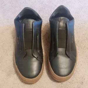 Svarta sneakers från Axel Arigato st 40 - Stilrena svarta sneakers från Axel Arigato i skinn med bruna gummisulor. Modellen har minimalistisk design utan snörning, med elastisk panel över vristen och diskret guldfärgad logga på sidan. Perfekta för dig som gillar clean och modern look.