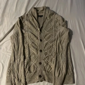 Beige stickad kofta från Red Herring - Snygg beige stickad kofta/cardigan från Red Herring med grovt kabelstickat mönster med bruna knappar. Koftan har långärmad design. 