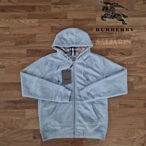 Grå Burberry Hoodie - Letar du efter en stilren och lyxig hoodie? Denna helt nya grå Burberry-hoodie kombinerar elegans med hög komfort. Den har Burberrys ikoniska rutmönster i fodret och en diskret logga på bröstet för en exklusiv touch. Tillverkad av mjukt, slitstarkt material och utrustad med dragkedja samt huva – både praktisk och stilfull.  📌 Skick: Ny med taggar. 📦 Snabb leverans – skickas samma dag! .