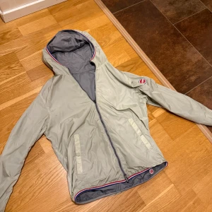 Colmar vändbar vindjacka med huva - Snygg vändbar vindjacka från Colmar med huva och dragkedja. Ena sidan är ljusbeige med röda och blå detaljer, andra sidan är grå. Jackan har två fickor på varje sida och Colmar-logga på ärmen. Perfekt för dig som vill ha två looks i en jacka.