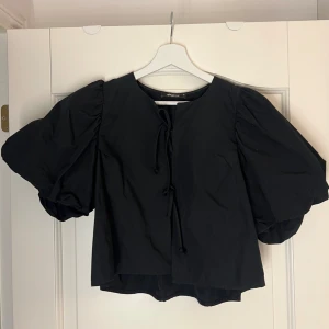 Svart blus med puffärmar och snörning - Supersnygg svart blus från Gina Tricot med puffiga korta ärmar och snörning framtill. Blusen har en rak passform och är tillverkad i ett mjukt bomullstyg. Perfekt för dig som vill ha en trendig och stilren look.