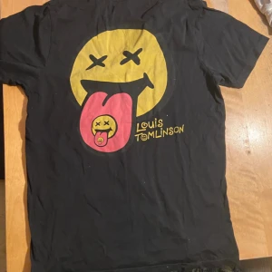 Louis Tomlinson svart t-shirt smiley - Svart t-shirt med stort tryck av en gul smiley med utsträckt rosa tunga på ryggen och mindre smiley på bröstet. Coolt grafiskt motiv och Louis Tomlinson-logga. Klassisk passform och rund hals. Perfekt för dig som gillar bandmerch och streetstyle.