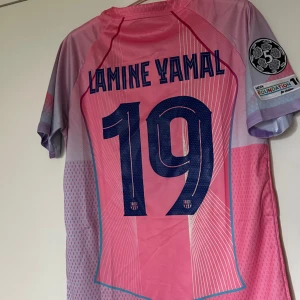 FC Barcelona Lamine Yamal  - Snygg FC Barcelona fotbollströja i rosa och ljuslila med Lamine Yamal och nummer 19 på ryggen. Tröjan har korta ärmar, klubbmärke, Nike-logga och Spotify som sponsor framtill. Tillverkad i lätt och ventilerande material, perfekt för match eller träning.
