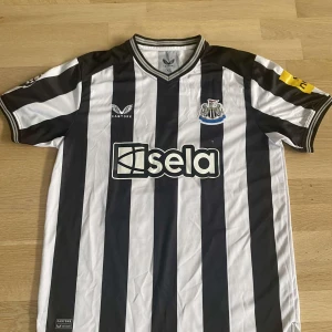 Newcastle United Isak 14 matchtröja XL - Newcastle Uniteds officiella matchtröja från Castore i storlek XL. Svart och vit med breda vertikala ränder, klubbmärke på bröstet, sponsorlogga 'sela' och Isak 14 tryckt i rött på ryggen. Klassisk V-ringning och korta ärmar. Materialet är lätt och andas. Pris går att diskuteras 
