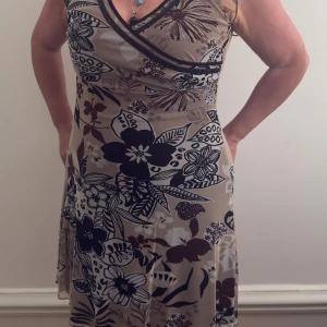 Blommig omlottklänning från Cue - Supersnygg kortklänning från Cue med omlott-ringning och blommigt mönster i svart, beige och brunt. Klänningen har ärmlös design och lätt utställd kjol, perfekt för sommarens alla tillfällen. Tunt och följsamt material som känns luftigt. Fungerar utmärkt till en S också.